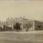 Image: a large stone public building with a low pitched roof situated on a street corner and fronting a park. The centre of the building is three-storeys high with the wings on either side at two storeys.