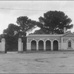 Image: Single-story stone building with a veranda featuring four archways. To the left of the building is a large metal gate with lights on top of the gate posts and a flag pole.