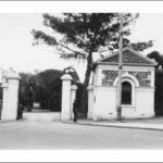 Image: Small stone building with a single arched window and gable roof next to open gates. A man stands next to the gates