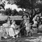 Image: Three women and seven children in 1950s clothing sit on low chairs in a park enjoying a picnic. In the background a rotunda can be seen.