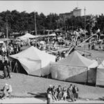 Image: a spinning carnival ride set up in a park is surrounded by a number of large canvas tents and a crowd of people in 1930s era clothing