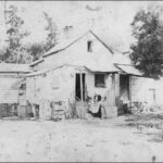 Image: A woman in a black dress and white apron stands on the stairs of a small 19th century weatherboard house