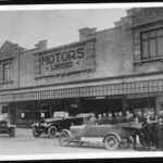 Image: a group of men in early 20th century clothing pose around a number of cars parked outside a two storey building with a sign reading "Motors Limited"