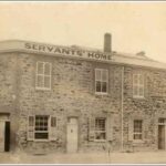 Image: A simple two-storey stone building with three main entrances, one on a corner, shuttered windows and a parapet sign reading "Servants' Home".