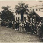 Image: A line of people in 1920s clothing stand next to the front of a white, two-storey building