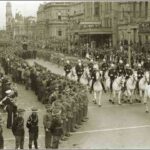 Image: a group of men in military dress uniform riding white horses parade past lines of soldiers and a crowd of civilians. Other groups marching under banners follow behind.