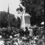 Image: crowd of people surrounding large bronze statue with cloth being removed.