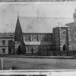 Image: a stone church with clerestory windows, a transept featuring a rose window, a square tower at the junction of the transept and nave and a arched porch at the front entrance.