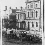 Image: horse drawn carriages are stopped on a dirt road outside of two public buildings. The one on the right is three stories while the one in the centre of the picture is two stories and features a columned portico with a decorative triangular pediment.