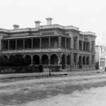 Image: a stone manse with a low pitched roof mostly hidden behind a parapet, an arched return verandah and columned balcony, and rectangular windows with triangular pediments.