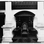 Image: a single chair sits on a small podium, located between two white columns and backed by carved wood panelling with a desk/shelf on either side the chair.