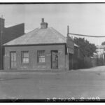 Image: a small semi-detached cottage with tin roof, rear lean-to and central chimney. Each residence has a door and single window on the front side of the house. A figure stands in the open door of one side of the building.