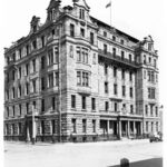 Image: an imposing six storey stone building with bay windows, a columned portico, balconies, mansard roof, curved parapets and a flagpole on the roof stands on the corner of two roads. A 1920s era car can be seen to the right of the photo..