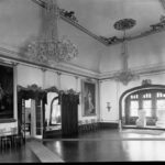 Image: A large, well-lit open room with wooden floors, crystal chandeleirs and large portraits hanging on its walls