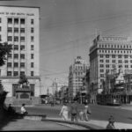 Image: a view along a city street lined with a number of tall public buildings of approximately ten stories interspersed with smaller two to four storey shops and hotels