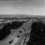 Image: electric trams and pedestrians travel down a wide city street which bisects a small city of mostly low lying buildings and passes through a large park