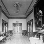 Image: An interior hallway with large painted portraits hanging from each wall and a double door at the end