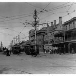 Image: electric trams travel down a wide city street lined with two and three storey buildings. A large, ornate lamp post can be seen in the centre of the image.