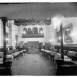 Image: leather bench seats sit behind desks facing inwards and arranged along the walls of a large room which is decorated with large portraits. At the far end of the room is a more ornate desk with three individual seats.