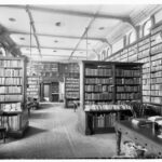 Image: a small library with leather bound volumes on tall wooden shelves. Most of the shelves line the walls but there are several rows protruding into the room from both sides. The room has a patterned carped and has desks and chairs scattered throughout