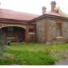 Image: A single storey bluestone building with red brick quoins, red tin roof with cream details and a number of brick chimneys. The building also has a verandah and decorative stained glass on two of its windows.