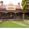 Image: A man and his dog stand outside a bluestone building with red brick quoins around its windows and two red brick chimneys. The building also has a red tin roof, simple wooden verandah and a gable with decorative moulding painted in cream and taupe.