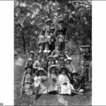 Image: A group of boys and girls, some holding sticks and flowers, with two young men