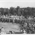 Image: a large crowd of people in early 20th century dress, some with umbrellas, stand on a grassed area in a park watching men in military uniform parade.