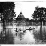 Image: A boy stands in waist deep water in a flooded park while others sit in row boats around him. In the centre of the photograph is a rotunda which is half underwater.