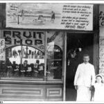 Image: A man and young girl in early 20th century clothes stand outside a fruit shop. The building is covered with advertising including that for icecream and cool drinks