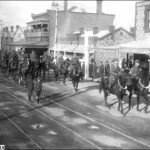 Image: a troop of mounted soldiers ride along a city street while women on the store balconies and two boys standing on the tram line watch on.