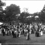 Image: a large group of people in 1920s fomalwear mingle on a lawn in front of a large building. A tent with white roof is visible at image left