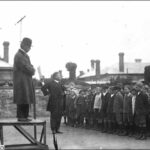 Image: a man in a coat and bowler hat stands on a small platform while below a headmaster in a dark suit addresses a group of boys and girls standing in neat rows.