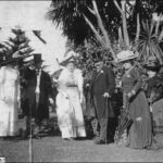 Image: A small group of well-dressed men and women stand on a lawn in front of trees