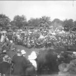 Image: a large crowd of people in 1920s dress, some with umbrellas, sit or stand in a park around a man on a low stage