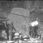 Image: Four men stand around a large piece of slate which is suspended by chains in a quarry