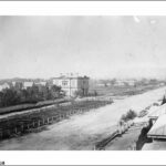 Image: A dirt street with a large, rectangular two-storey building and other smaller structures