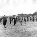 Image: a group of boys and girls in early 20th century clothing and hats stand in lines in a field with their arms outstretched to either side.