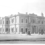 Image: A large, two-storey building fronted by a dirt street and two horse-drawn carts