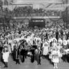 Image: A large group of women and children wearing flower costumes stand in an large open room with a sign reading English Dandelion while in the gallery above them men and women look on