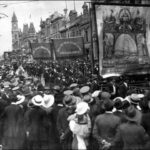 Image: men in dark suits parade under banners past a large crowd