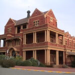 Image: Modern photograph of a large red brick building with yellow painted decorative details such as columns, and featuring a verandah and balconies.