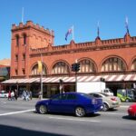 Image: A large brick building with archway windows and a square turret at one end. Cars drive by on a busy street in the foreground