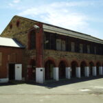 Image: a two storey stone building with a brick arched loggia and barred upper storey balcony. A security camera is mounted on one end of the building.