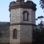 Image: A two-storey octagonal stone tower set within a high wall with a crenellated parapet.