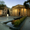 Image: The front of a large stone building with six columns flanking the entrance. A fountain and benches are arranged along stone sidewalks in front of the building