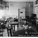 Image: Black and white image of the Peterborough room inside the Cheer up hut. The image shows a piano in the right far corner and a semi circular seat in the far left corner. Also in the room are dining settings and lounges.