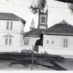 Image: street view of a white stone mansion with an additional building adjoined and two church towers in the background.