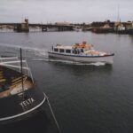 Image: A small motor boat travels down a river past a larger ship. A drawbridge is visible in the background