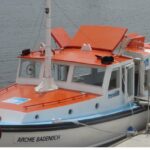 Image: A small motor boat is moored alongside a dock. It is painted white with an orange foredeck and blue gunwales